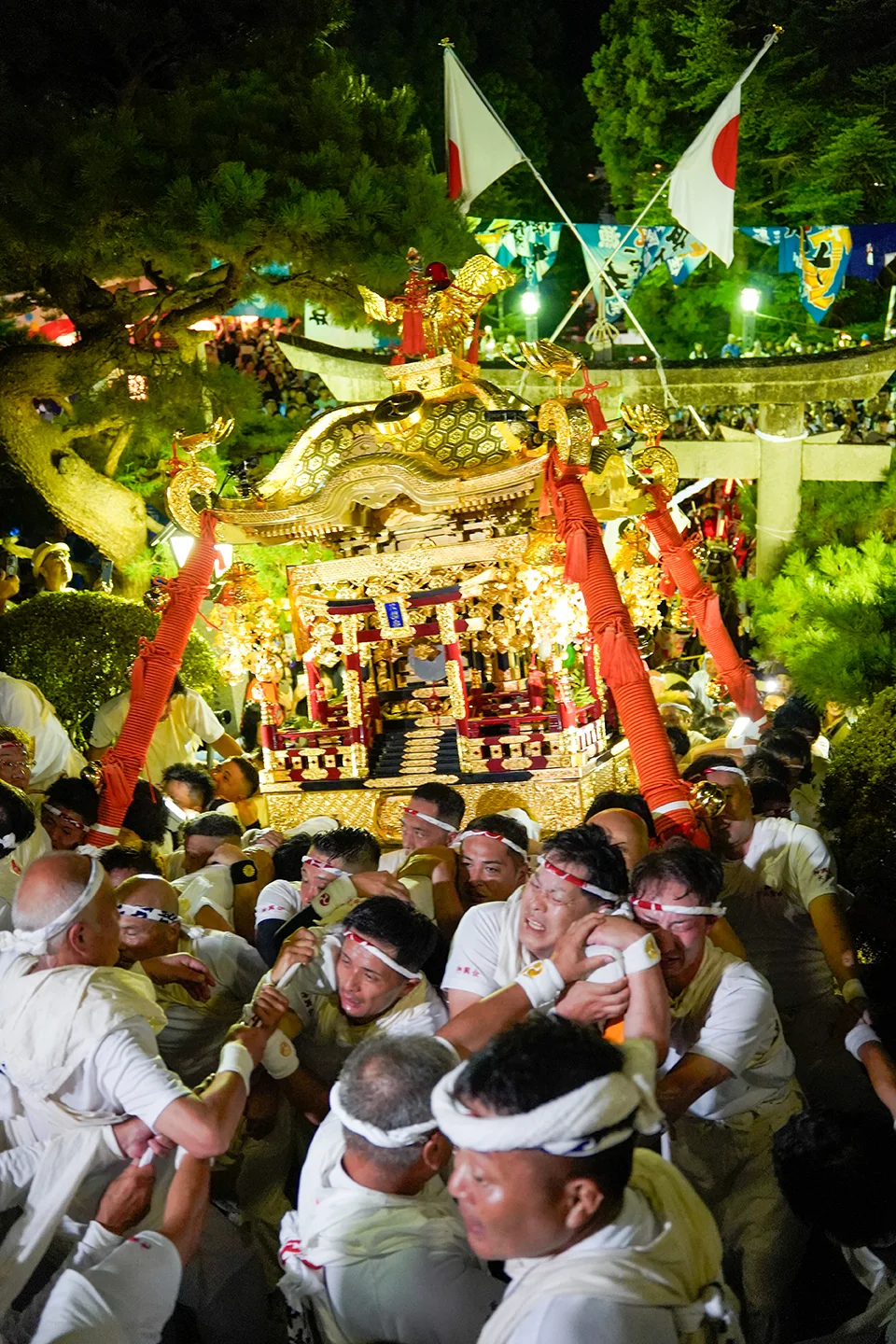 White-clad men jostling under a torii gate carrying a golden portable shrine at night during Yamada Matsuri