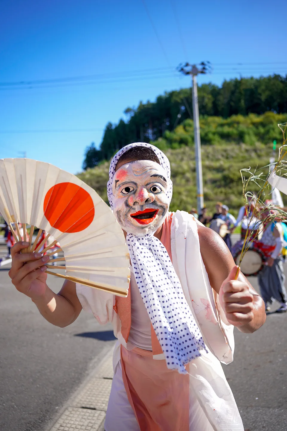 Masked comic performer dancing with a rising-sun fan at Yamada Matsuri